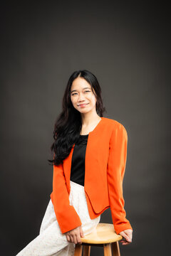 Portrait of a smiling young Southeast Asian Malaysian woman wearing professional corporate attire, featuring a bright orange blazer and black top with a confident pose