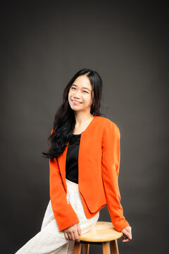 Portrait of a smiling young Southeast Asian Malaysian woman wearing professional corporate attire, featuring a bright orange blazer and black top with a confident pose