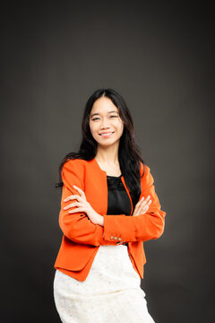 Portrait of a smiling young Southeast Asian Malaysian woman wearing professional corporate attire, featuring a bright orange blazer and black top with a confident pose