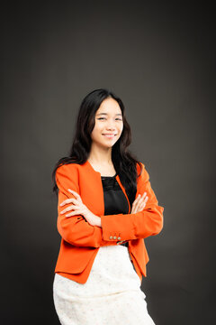 Portrait of a smiling young Southeast Asian Malaysian woman wearing professional corporate attire, featuring a bright orange blazer and black top with a confident pose