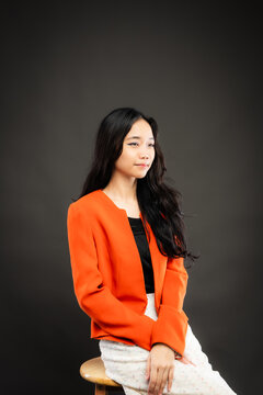 Portrait of a smiling young Southeast Asian Malaysian woman wearing professional corporate attire, featuring a bright orange blazer and black top with a confident pose