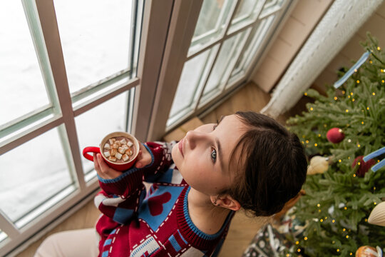 Young woman looking out snowy window with hot chocolate