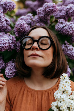 Woman smelling purple lilac flowers with closed eyes, wearing thick round black glasses, freckled face enjoying nature spring fragrance in terracotta dress, peaceful April portrait in blooming garden