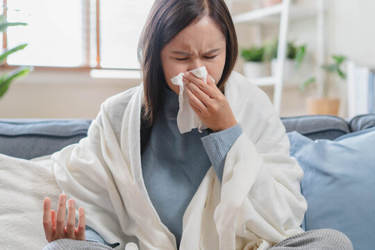 Sick Asian woman blowing her nose with tissue on sofa, wrapped in blanket, suffering from high fever, seasonal flu, influenza virus infection, and cold symptoms at home