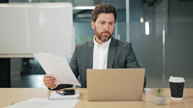 Professional immersed in work, reviewing important business documents at modern office desk. Engaged with laptop and papers, focused on critical information.