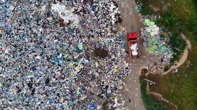 High angle drone view of a red garbage truck among huge piles of plastic trash and household waste at an open-air landfill site.
