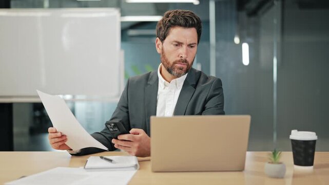 Focused business professional reviews documents, uses smartphone at office desk. Laptop, coffee present. Productive work environment for busy executive.