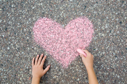 Little child girl hand fingers holding chalk and drawing pink heart shape on dark gray asphalt background in summer day. Closeup. Point of view shot. Top down view.
