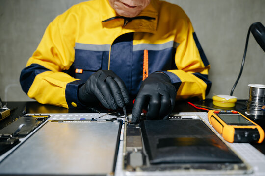 Senior Asian technician using tweezers to repair a disassembled tablet on a workbench in a modern service workshop.