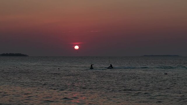 An aerial drone view of the sunset over the waters off Tanjung Gelam Beach, Karimunjawa Island. Silhouettes of tourists riding jet skis are visible.