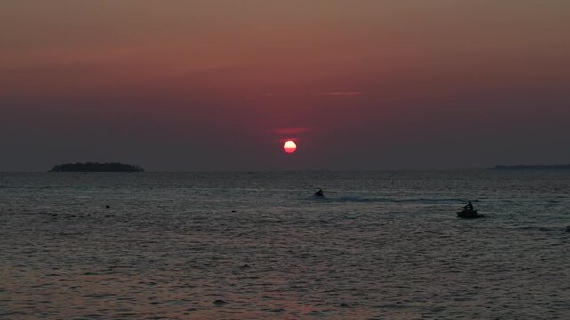 An aerial drone view of the sunset over the waters off Tanjung Gelam Beach, Karimunjawa Island. Silhouettes of tourists riding jet skis are visible.