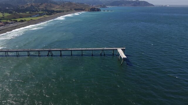 Aerial drone view showing the Pacifica Municipal Pier jutting out into the deep blue Pacific Ocean, with the scenic California coastline and sandy beach stretching into the distance