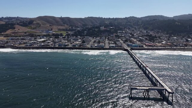Aerial perspective of the historic Pacifica Municipal Pier stretching into the glistening Pacific Ocean, with the coastal cityscape and rolling hills of Northern California in the background