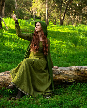 full length portrait of beautiful female model, wearing green medieval historical gown costume with cape. sitting figure pose on tree branch.  Magical lake forest background 