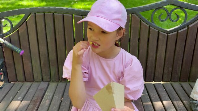 Young European girl in pink hat eating french fries on a park bench