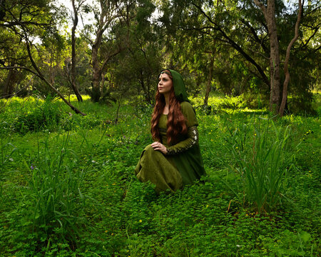 full length portrait of beautiful female model, sitting  kneeling figure picking herb plants, wearing hooded green medieval historical gown costume with cloak. Magical forest background with trees.