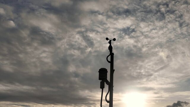 Anemometer Under Cloudy Sky: A detailed shot of an anemometer on a pole, capturing the atmospheric conditions under a vast, dramatic sky filled with textured clouds and the soft glow of the sun.