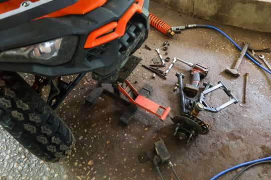 Off road vehicle front end with removed wheel and scattered mechanical tools on dirty workshop floor, gritty repair scene showing suspension parts and pneumatic hose