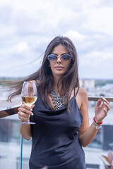 Fototapeta premium Beautiful fashionable woman holding a glass of white wine on a balcony