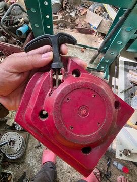 Red recoil starter housing held in hand with pull cord handle, dusty plastic cover showing screw holes and wear, workshop tools and shelving background with scattered parts and concrete floor