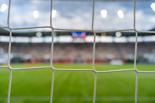 Soccer goal net in the foreground with a blurred football field, stadium and spectators in the background, representing competition, sport and victory.