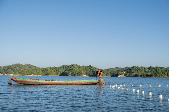 Rangamati, Bangladesh - 14 December 2024: View of a fisherman casting a net from a wooden boat on the calm waters of Kaptai Lake, surrounded by lush green hills under a clear blue sky.