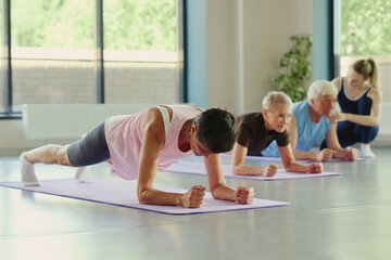 Fototapeta premium Mature Caucasian woman performing forearm plank on yoga mat during fitness class with senior people. Image represents active aging and is suitable for wellness marketing