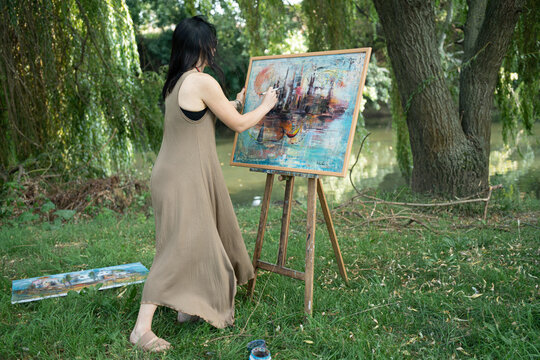 Woman artist painter outdoor painting a canvas on an easel from behind by the river, rear view