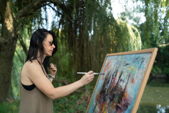Beautiful woman painter by the river with sunglasses painting on a canvas on an easel