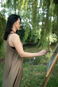 Beautiful woman painter by the river with sunglasses painting on a canvas on an easel