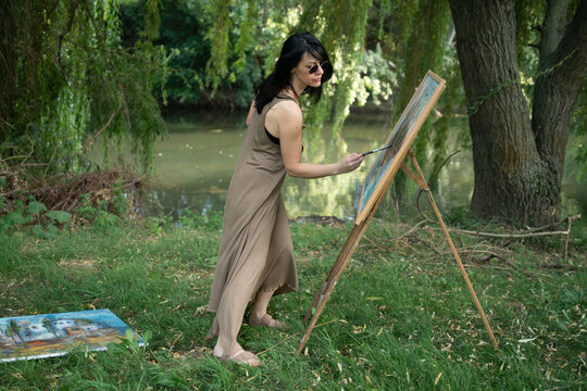 Beautiful woman painter by the river with sunglasses painting on a canvas on an easel