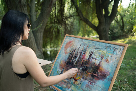 Beautiful woman painter by the river with sunglasses painting on a canvas on an easel, behind view over the shoulder