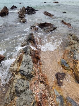High angle top-down view looking into a narrow sandy crevice surrounded by barnacle-covered coastal rocks and clear incoming sea water.