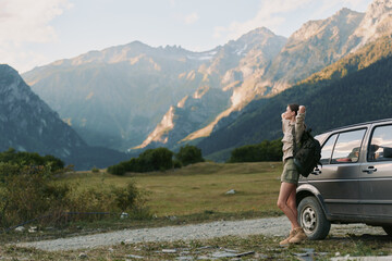 Obraz premium A lone young woman with a backpack stands beside a parked car in a rugged mountain valley, preparing for a hike and enjoying a moment of solitude. A feeling of independence.