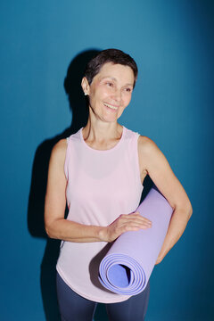 Smiling senior Caucasian woman holding yoga mat against blue background, promoting active aging and wellness. Ideal for senior fitness and healthy lifestyle marketing, vertical