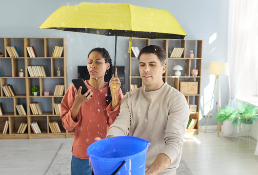 Couple woman man manage water leak with umbrella. Indoors they catch drips in a bucket as the woman uses phone to call a plumber for fast repair. Clear concept of urgent home leak emergency.