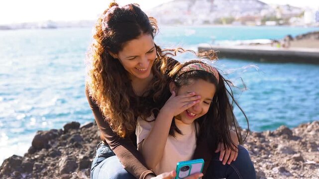 Happy mother and daughter using smartphone on the beach during summer vacation - Family lifestyle, bonding and technology concept	