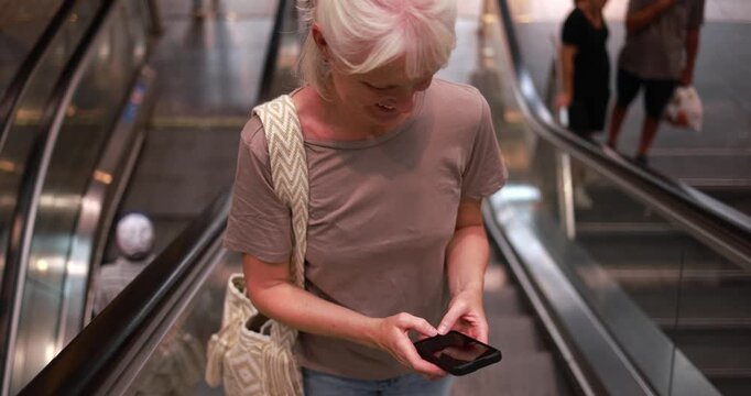 Caucasian senior woman using smartphone on metro escalator - City lifestyle, travel and technology concept - UGC content