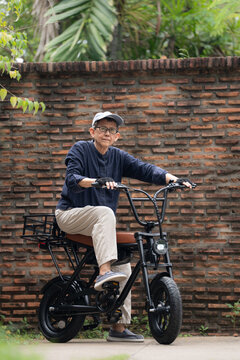 Elderly Gay Woman on Electric Bicycle: Senior LGBT woman wearing cap and gloves enjoys a ride on an e-bike, pausing to look out into the distance, with a brick wall as backdrop. Senior Gay woman
