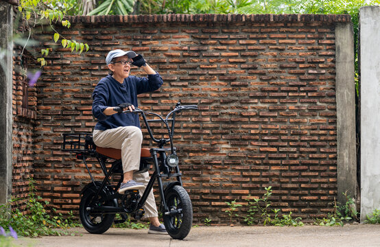 Elderly Gay Woman on Electric Bicycle: Senior LGBT woman wearing cap and gloves enjoys a ride on an e-bike, pausing to look out into the distance, with a brick wall as backdrop. Senior Gay woman