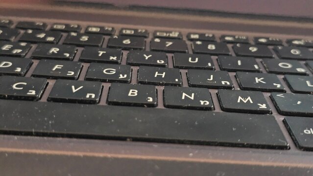 A close-up macro shot of a dusty black laptop keyboard with bilingual Latin and Hebrew character keycaps.