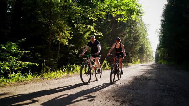 Two cyclists ride side by side along a sunny forest dirt road, enjoying outdoor exercise, nature, and peaceful adventure in a lush green woodland setting