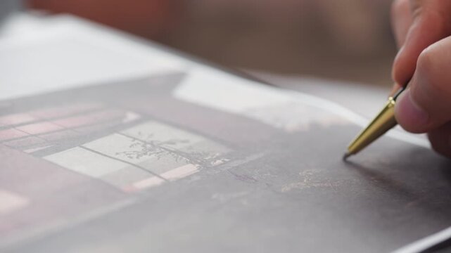 Close-up shot showing a hand holding a gold-tipped pen, tracing or pointing on a sheet with graphic printing