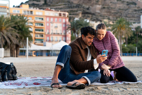 Couple relaxing on winter beach, scrolling social media on smartphone