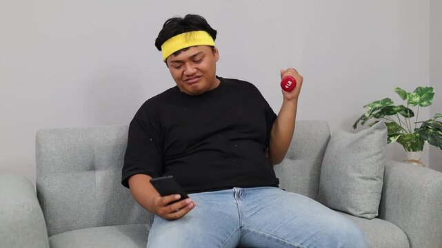 Asian Overweight man on couch with phone, dumbbell workout. Checking fitness tips, Lazy home workout procrastination concept theme.