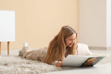 Fototapeta premium Little girl reading book while lying on floor at home