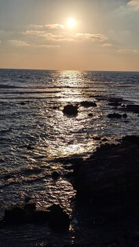 The sun reflects a golden path across calm ocean water near a rocky coastline at sunset.