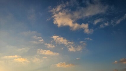 Scattered white and golden-tinted cumulus clouds drift across a deep blue sky during golden hour light. © Noa Badel