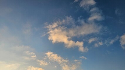 Scattered white and golden-tinted cumulus clouds drift across a bright blue sky during golden hour lighting. © Noa Badel