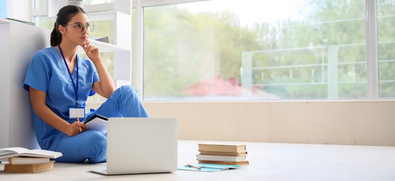 Thoughtful female medical student in blue scrubs and glasses studying on the floor with laptop and books, She is surrounded by books and a laptop, reflecting on her studies